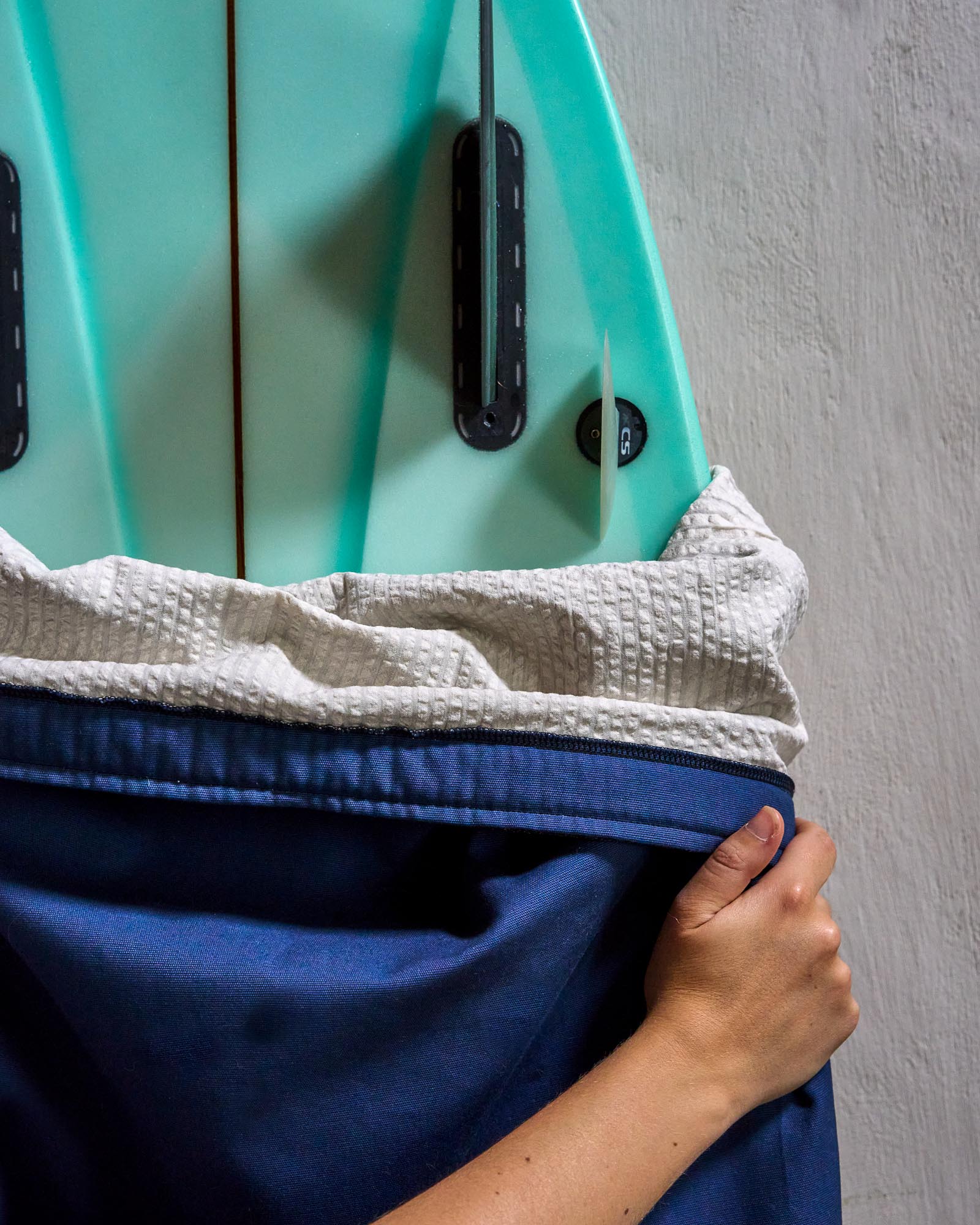 Person putting a blue surfboard cover on a turquoise surfboard against a grey wall.