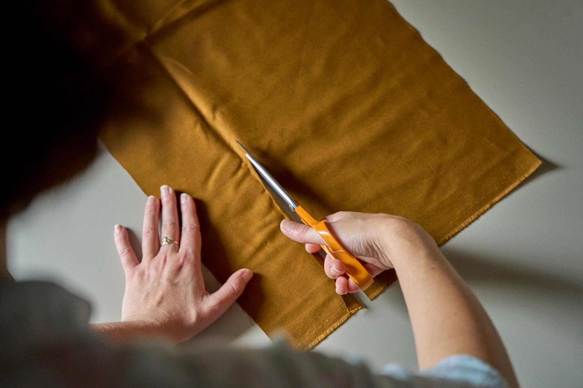 Person cutting brown fabric with orange scissors on a grey desk.