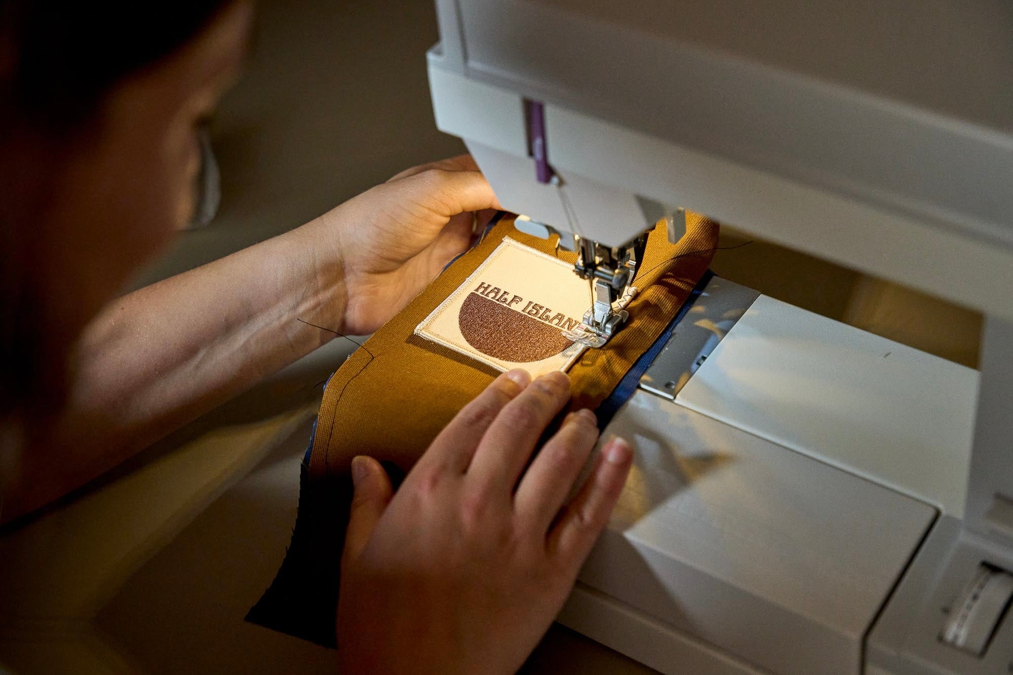 Person using a sewing machine to stitch a Half Island logo to some fabric.