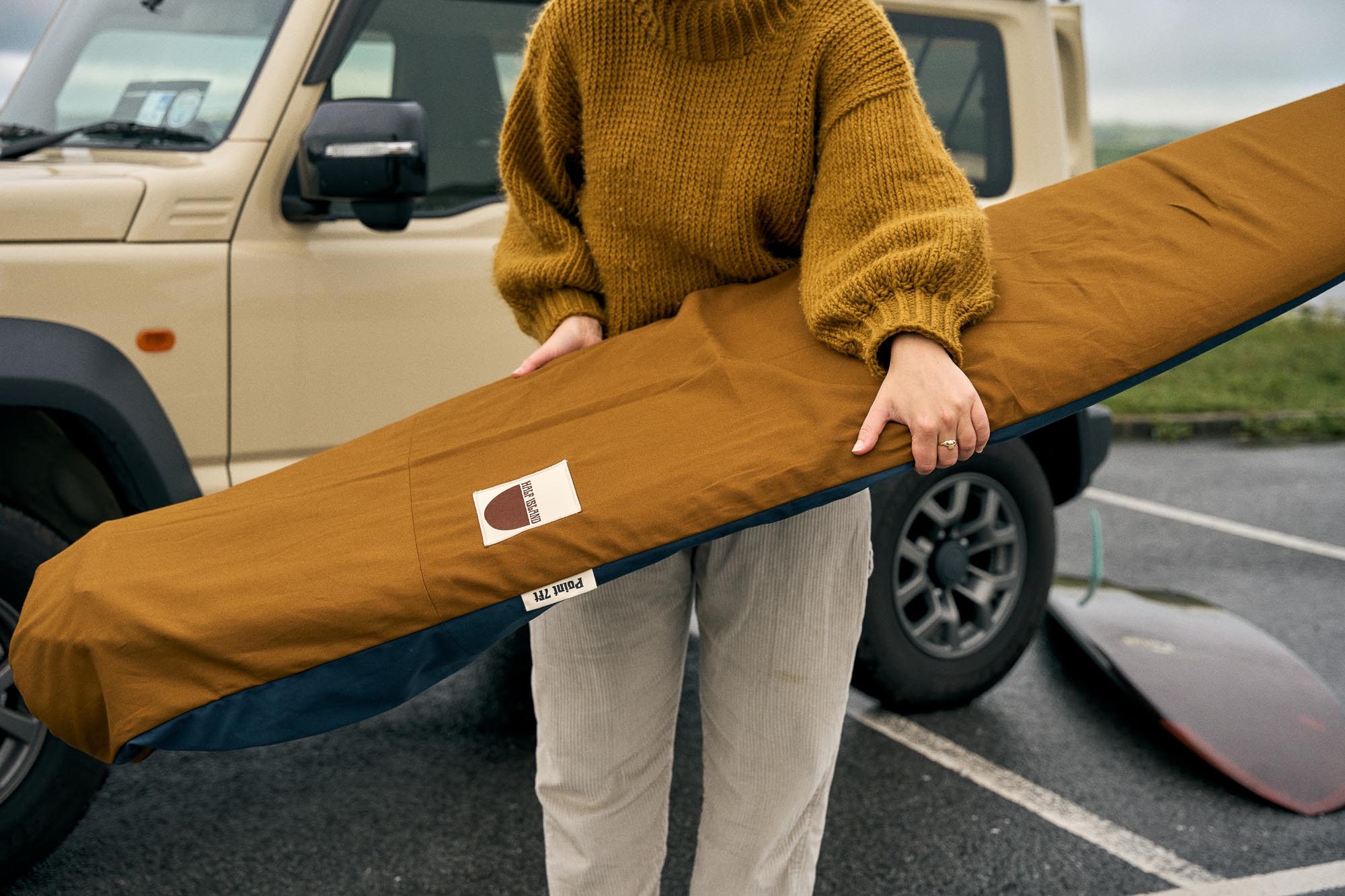 Person holding a surfboard in a brown board bag with a vehicle in the background.