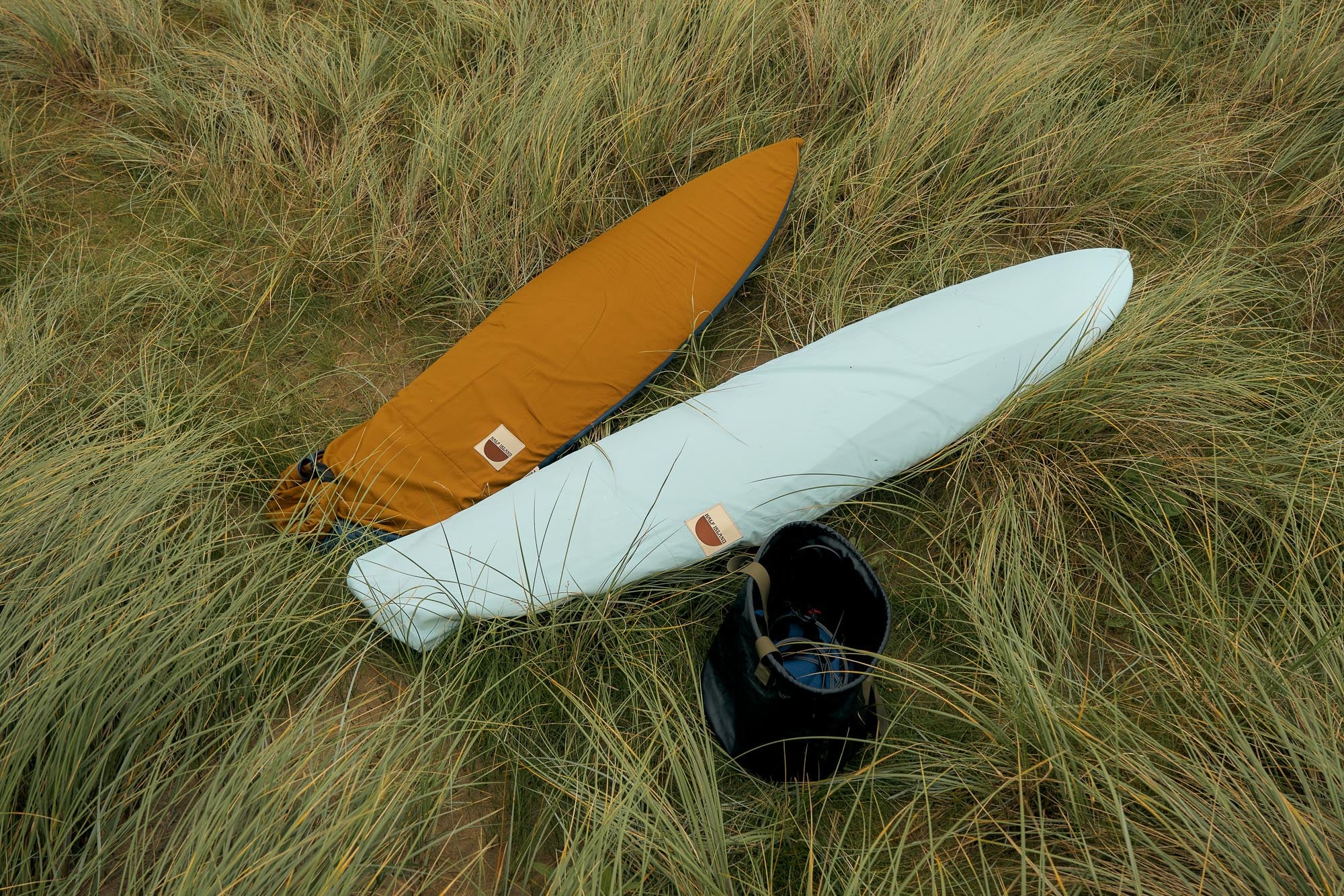 Two surfboards, one in a brown board cover and one in a light blue board cover, lying on grassy dune.