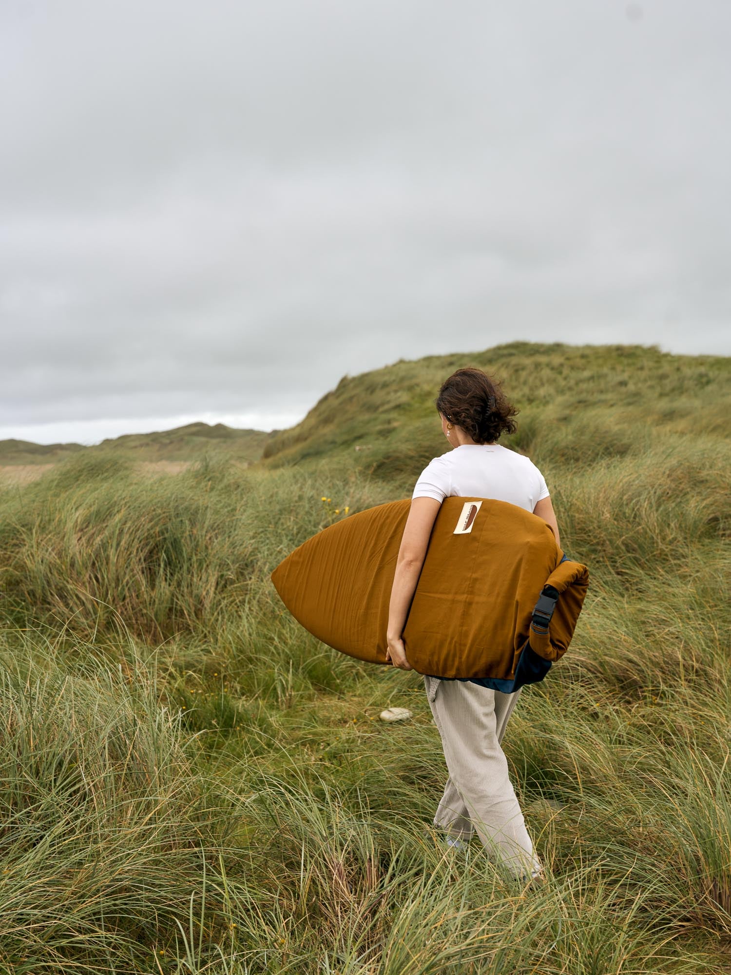 Person carrying a surfboard in a brown board bag through grassy dunes on a cloudy day.