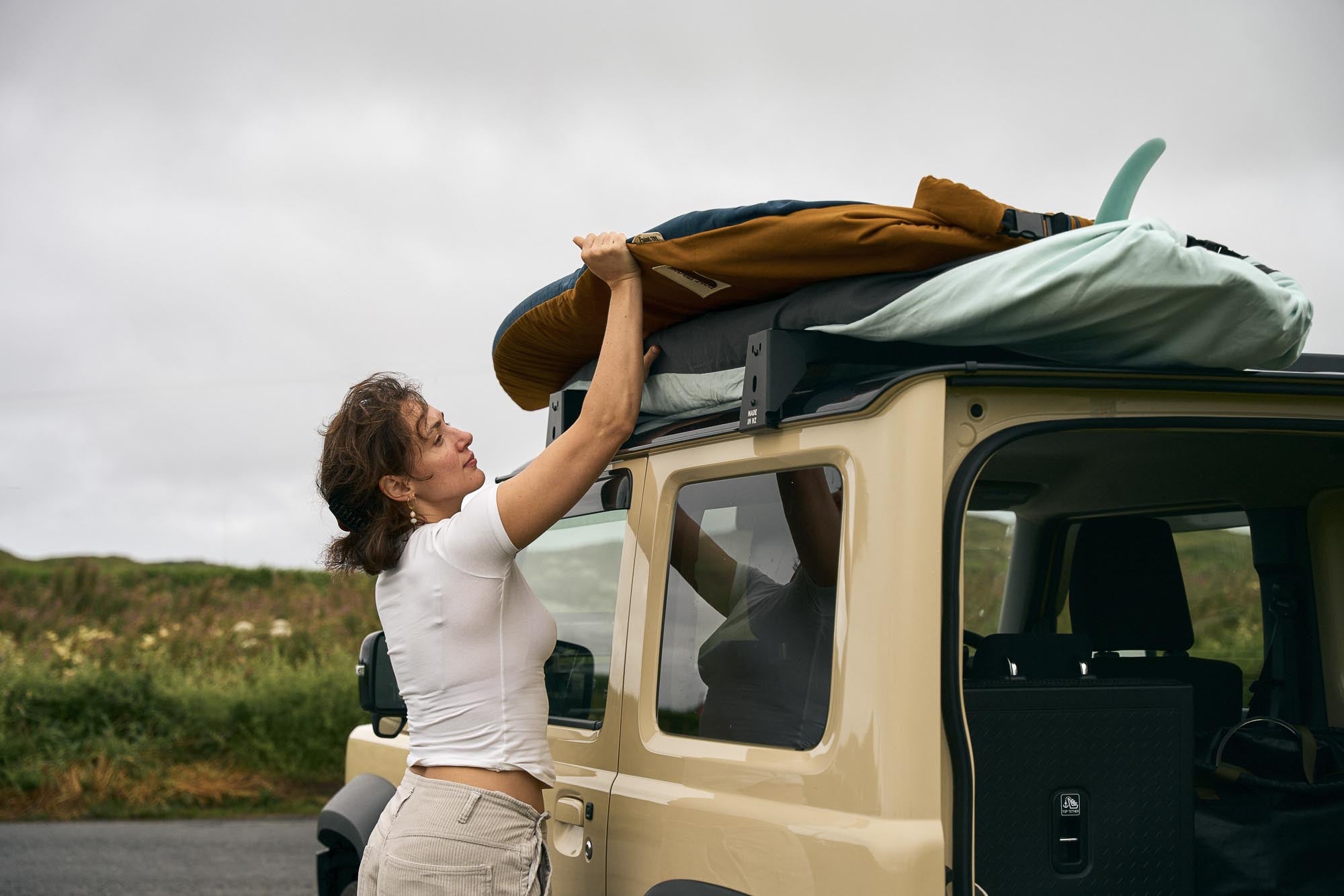 Person loading a surfboard onto a car roof with a scenic background.
