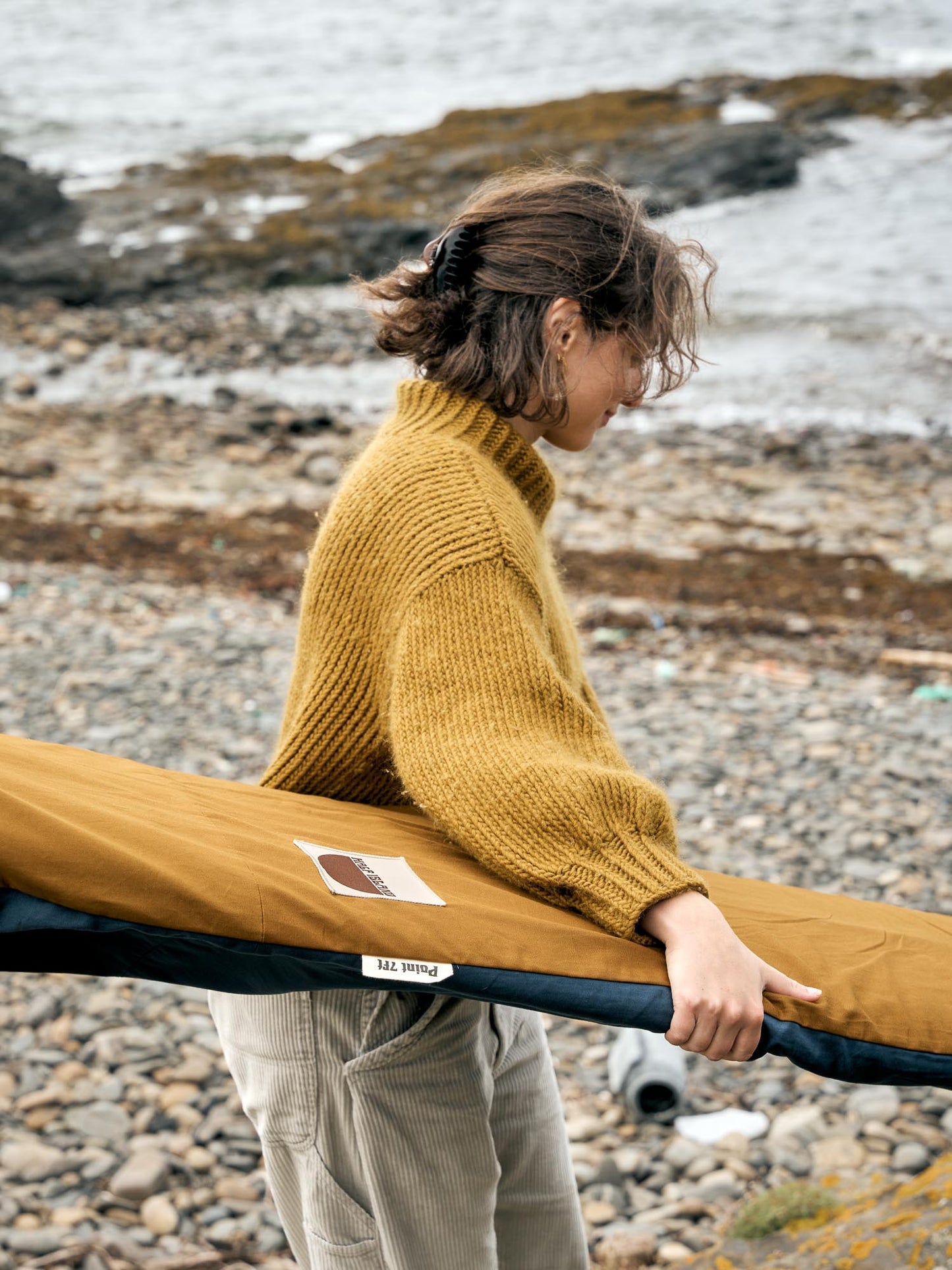 Person holding a surfboard in a brown surfboard cover on a rocky beach.