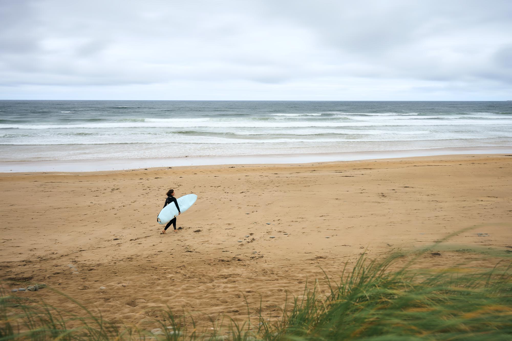 Person walking on a beach with a surfboard under an overcast sky.