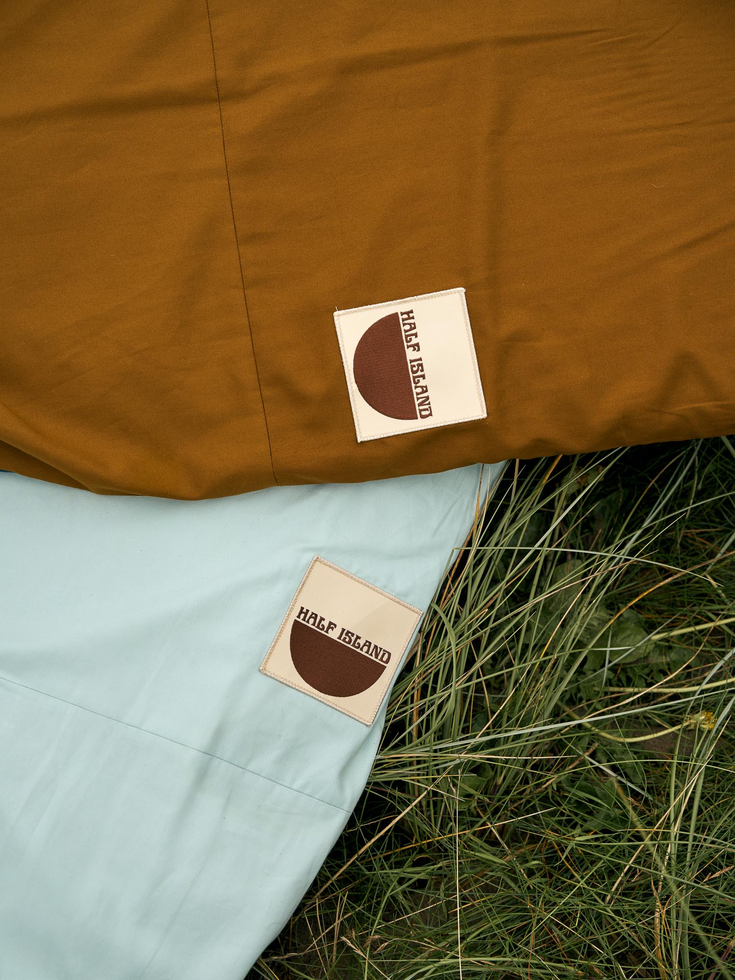Two fabric surfboard in board bags , one brown and one light blue, with 'Half Islands' logos on grass.