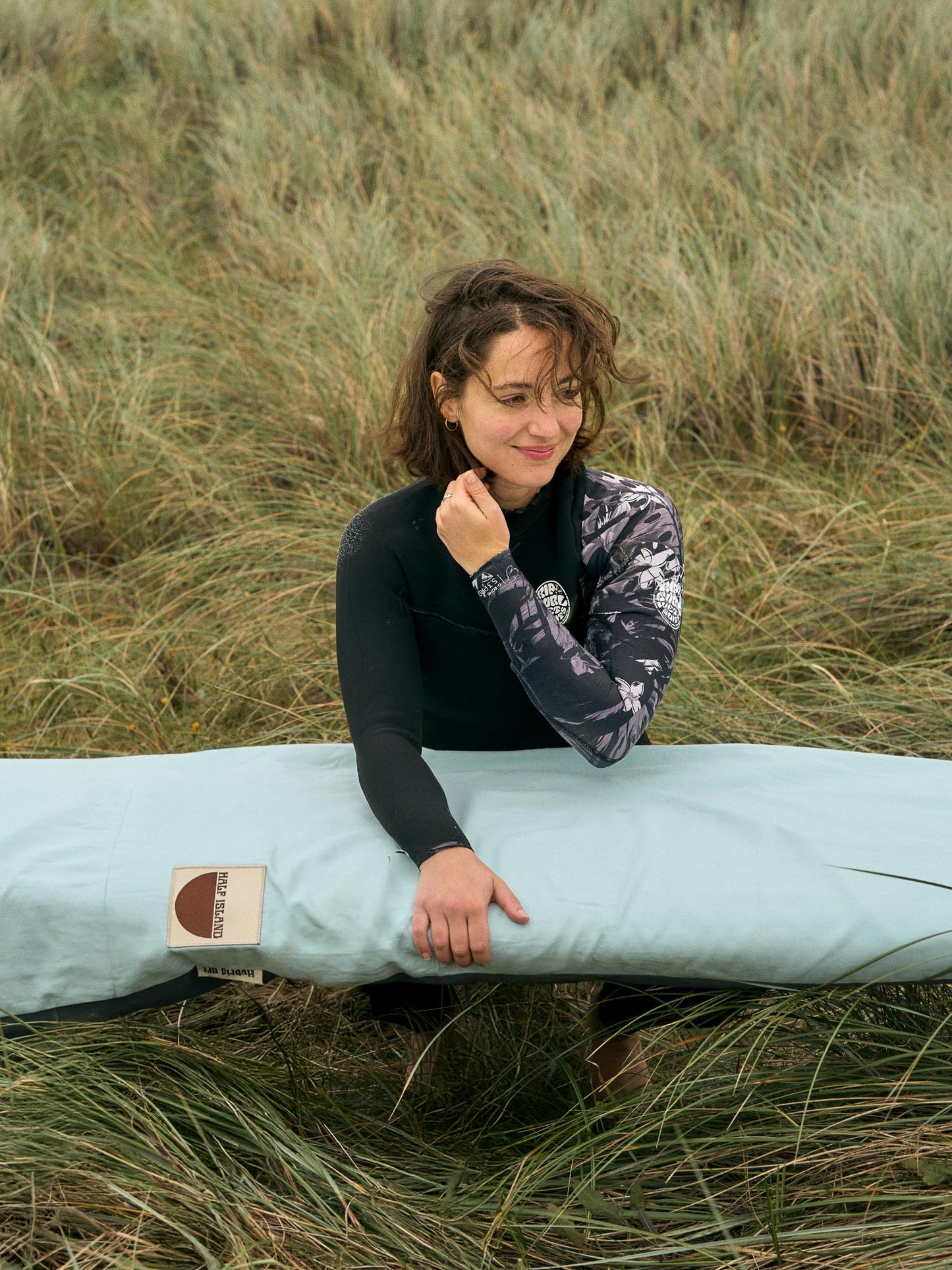 Person holding a surfboard in a light blue bag with a logo sitting in a grassy dune.