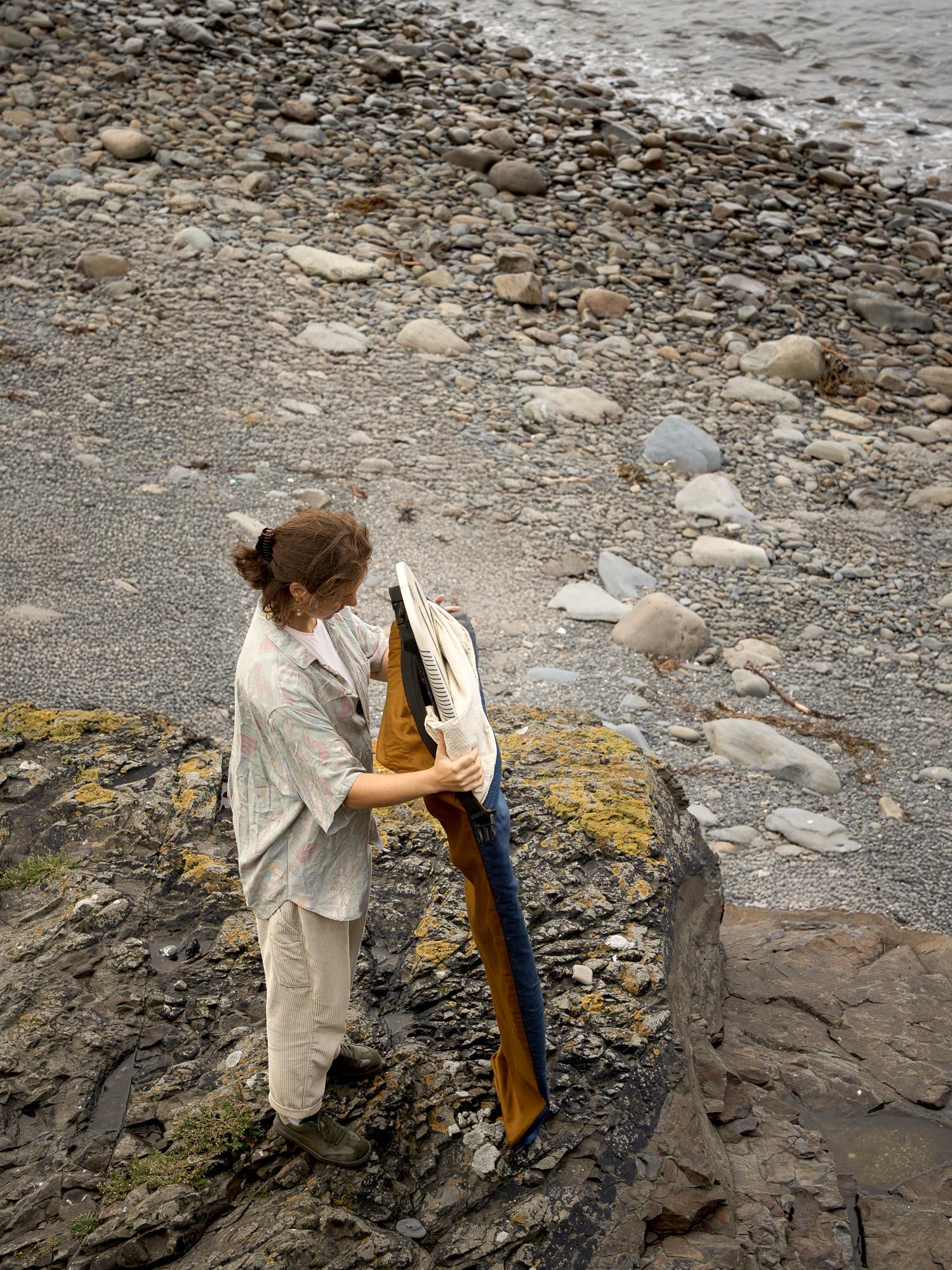 Person putting a brown board cover on a white surfboard on a rocky beach.