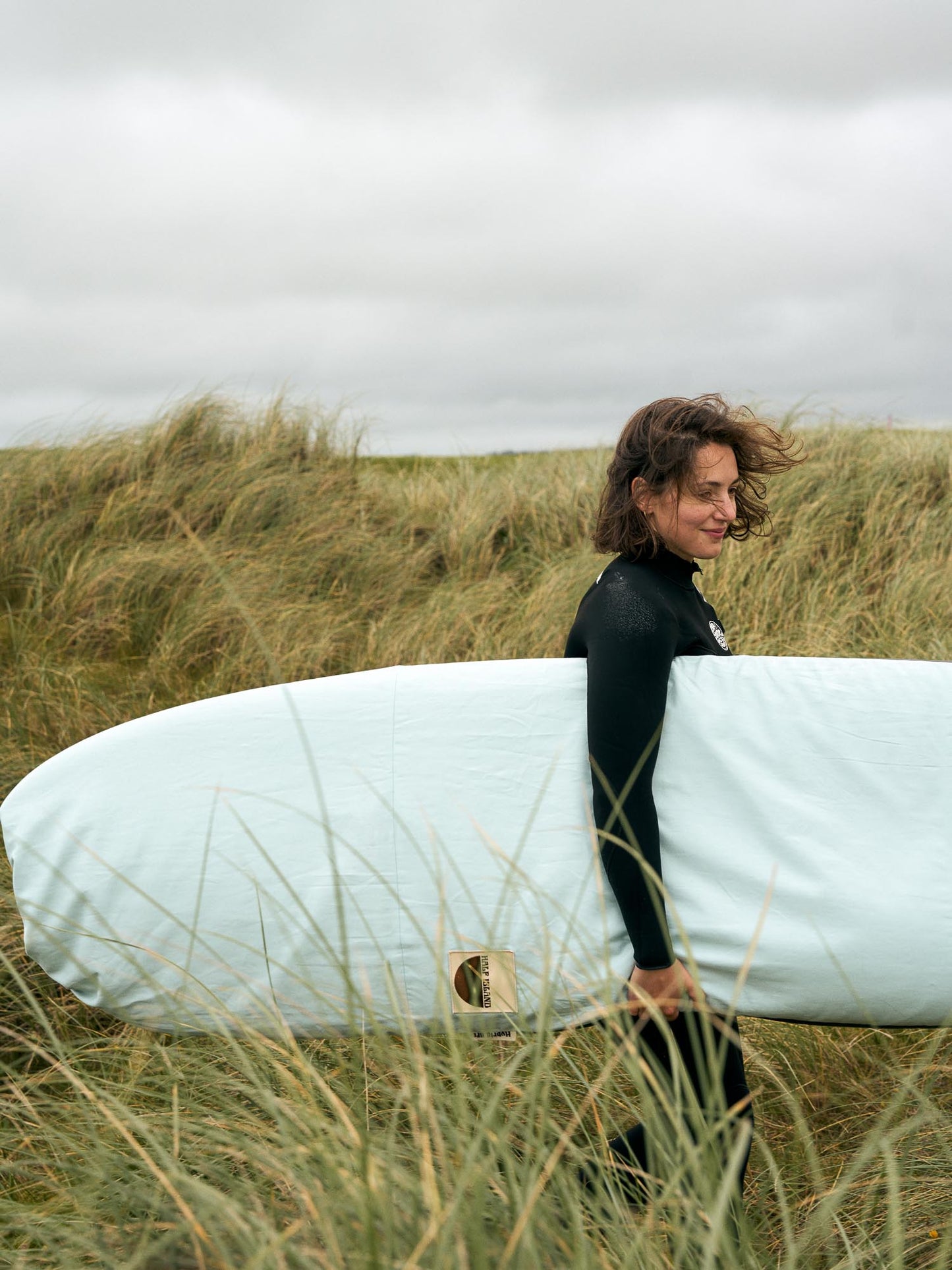 Person in a wetsuit holding a surfboard in a light blue board bag walking through sand dunes.