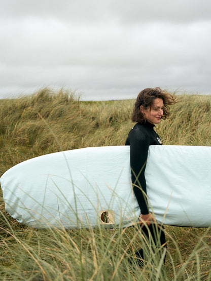 Person in a wetsuit holding a surfboard in a light blue board bag walking through sand dunes.