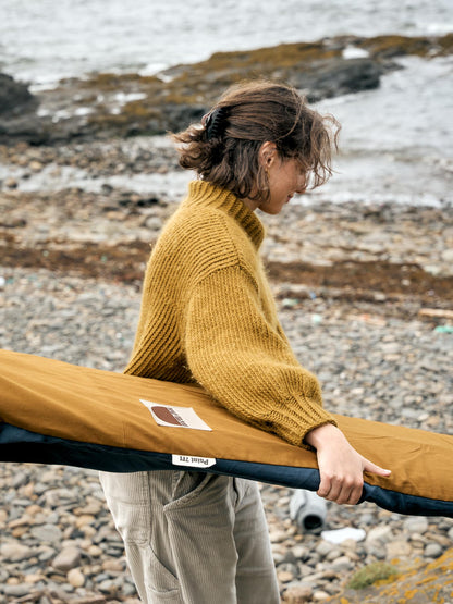 Person holding a surfboard in a brown surfboard cover on a rocky beach.