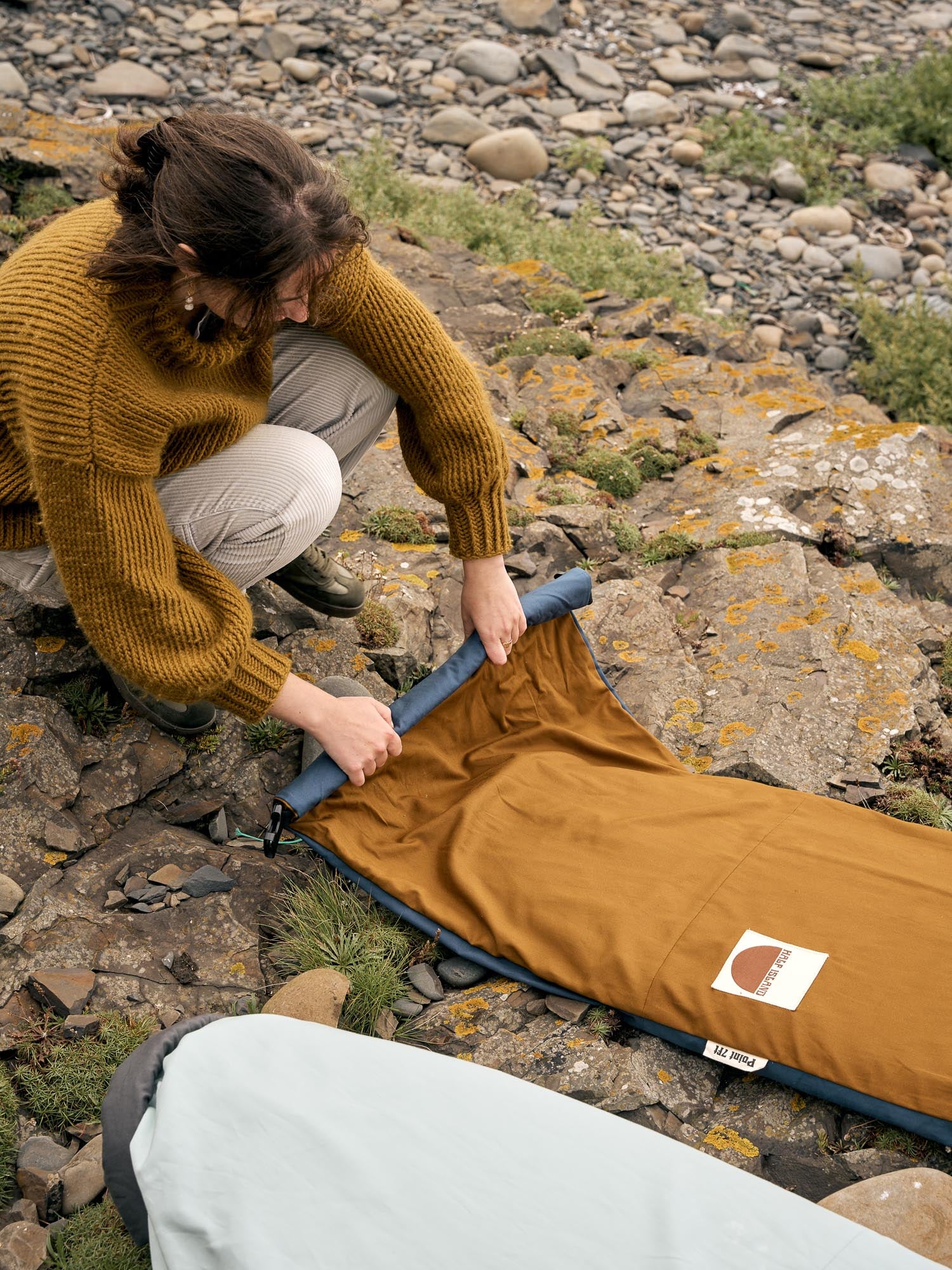 Person rolling up a brown board bag on a rocky shore.