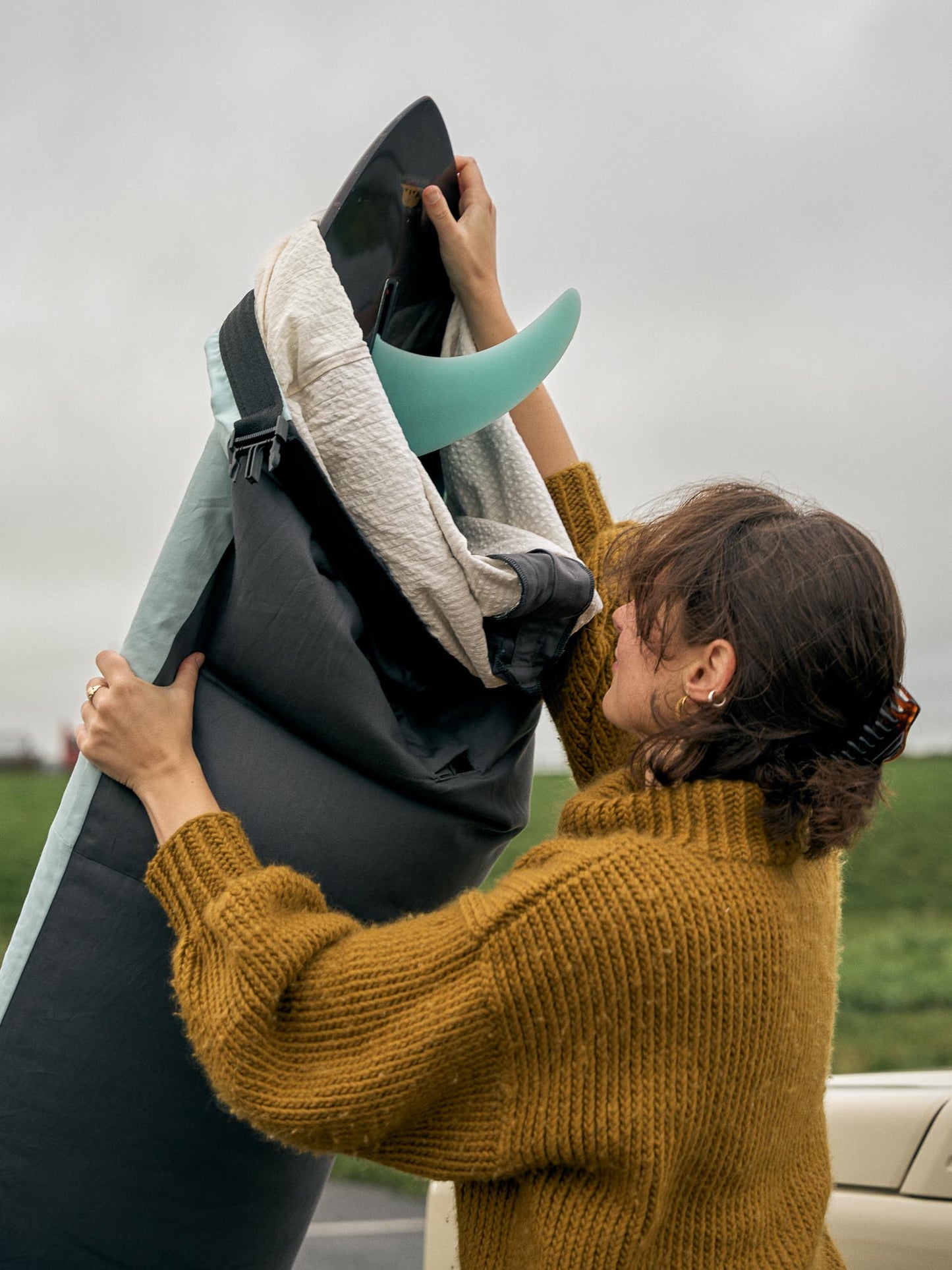 Person in a taking a grey surfboard bag of a single fin surfboard against a cloudy sky.