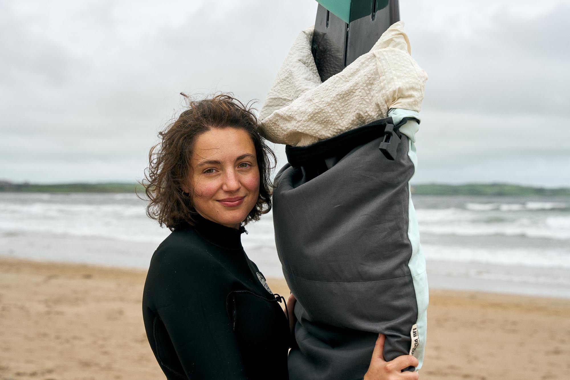 Person holding a surfboard with a grey surfboard bag with a sandy beach and waves in the background.